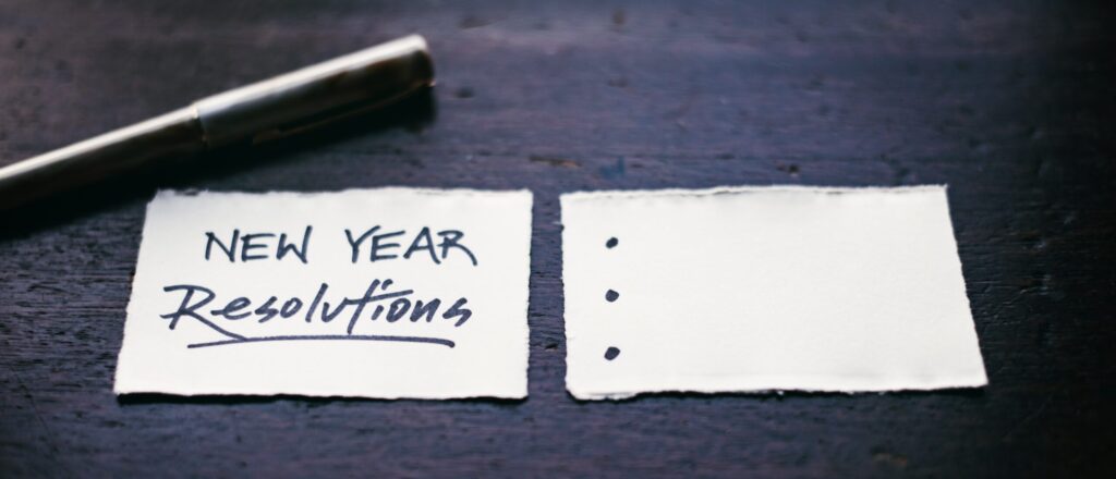 Two pieces of paper on a table with a pen and written on one paper is "New Year Resolutions".