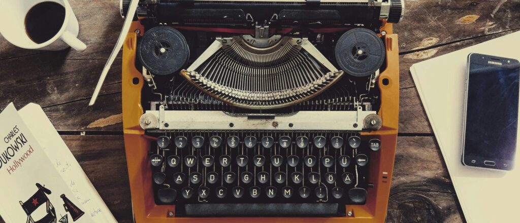 A typewriter on a wooden table, next to a book, cellphone, and a cup of coffee.